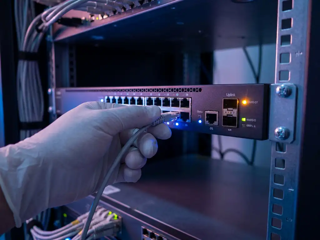 Technicianshand die een beheerkabel aansluit op een rackmount fiber switch in een donkere serverrack met amber waarschuwingslichten.