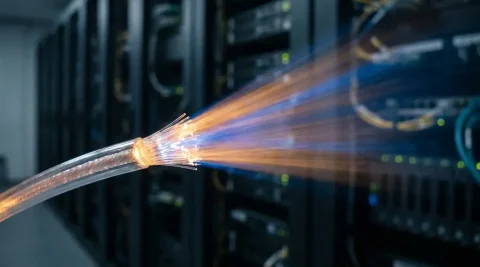 Gloeiende glasvezelkabel die licht breekt in een blauw-amber straal in een donkere serverruimte, met zichtbare glaskerntextuur.