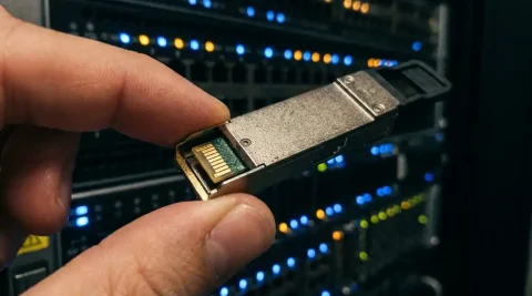 QSFP transceiver module tussen vingers gehouden boven een high-density fiber patch panel met knipperende poort-LED's op de achtergrond.