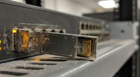 Verouderde SFP-transceiver half uit enterprise switch-poort getrokken, met geoxideerde goudkleurige contacten op grijs serverrek-schap.