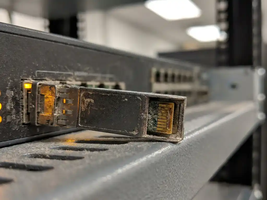 Verouderde SFP-transceiver half uit enterprise switch-poort getrokken, met geoxideerde goudkleurige contacten op grijs serverrek-schap.