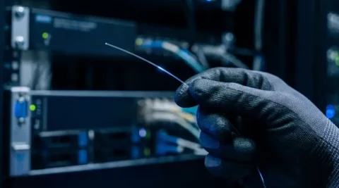 Gehandschoende hand houdt een glasvezelkabel omhoog in blauw licht, met serverrack op de achtergrond.
