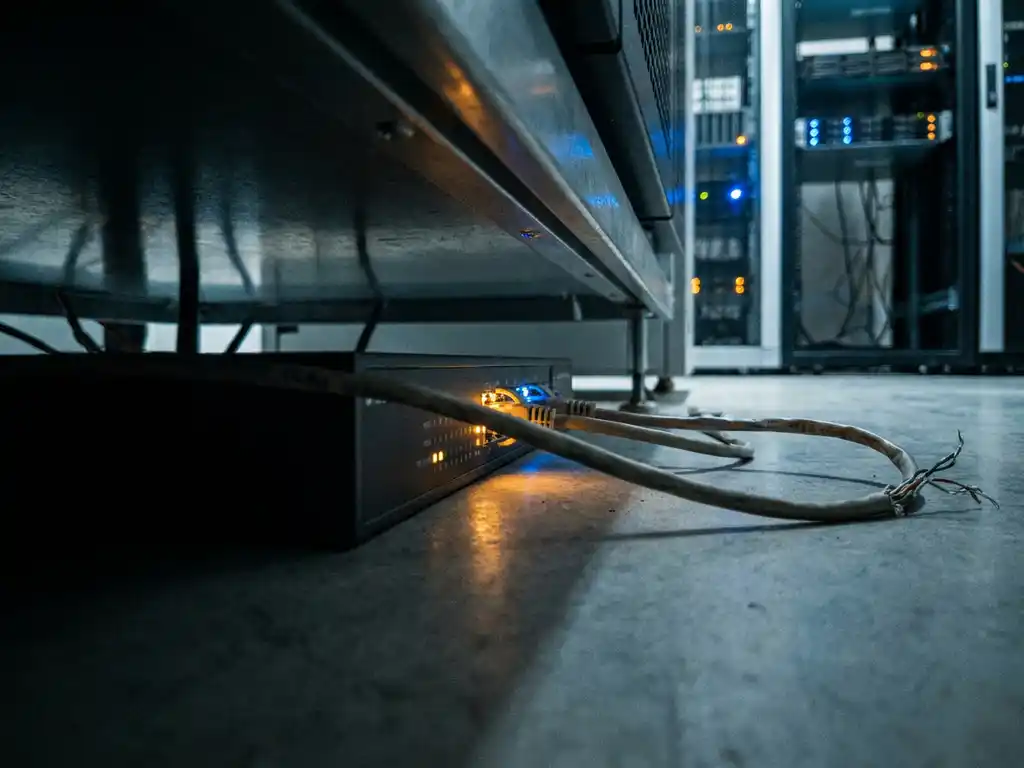 Enterprise netwerkrouter op serverruimtevloer met flikkerende statusindicatoren, loshangende ethernet-kabel en serverracks op achtergrond.
